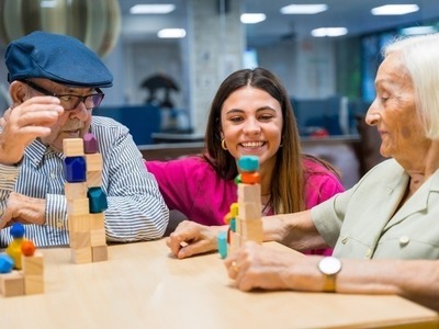 Image of two older people sitting at a table enjoying an activity involving wooden blocks whilst a support worker looks on and smiles.