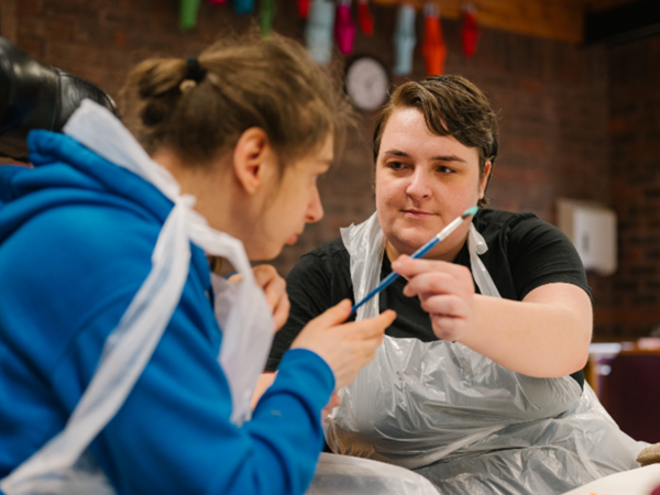 An adult social care professional handing a paint brush to an adult social care service user during a painting activity.